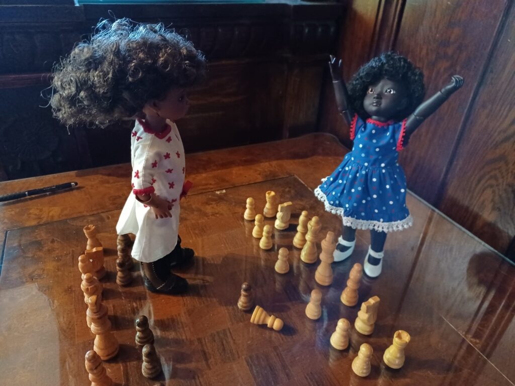 Two dolls standing on a chess table among chess pieces.