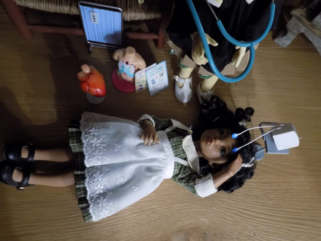 A doll and other figurines lines up on a tabletop with toy medical instruments.
