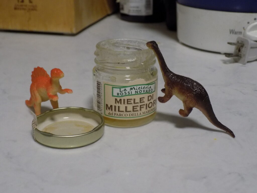 A jar of honey surrounded by toy dinosaurs.