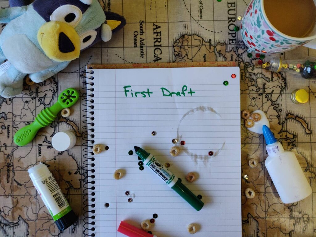 A lined notepad with the words “First Draft” sits on a kitchen table, surrounded by scattered sequins, glue, teething toys, cereal, and a half-drunk cup of tea. A stuffed animal of Bluey peeks in from the side.
