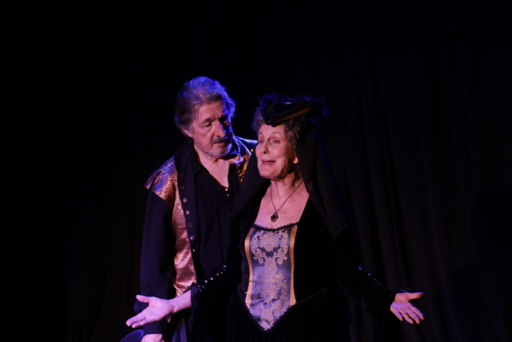 Two characters standing on the stage, delivering lines. Black background and mid-shot.