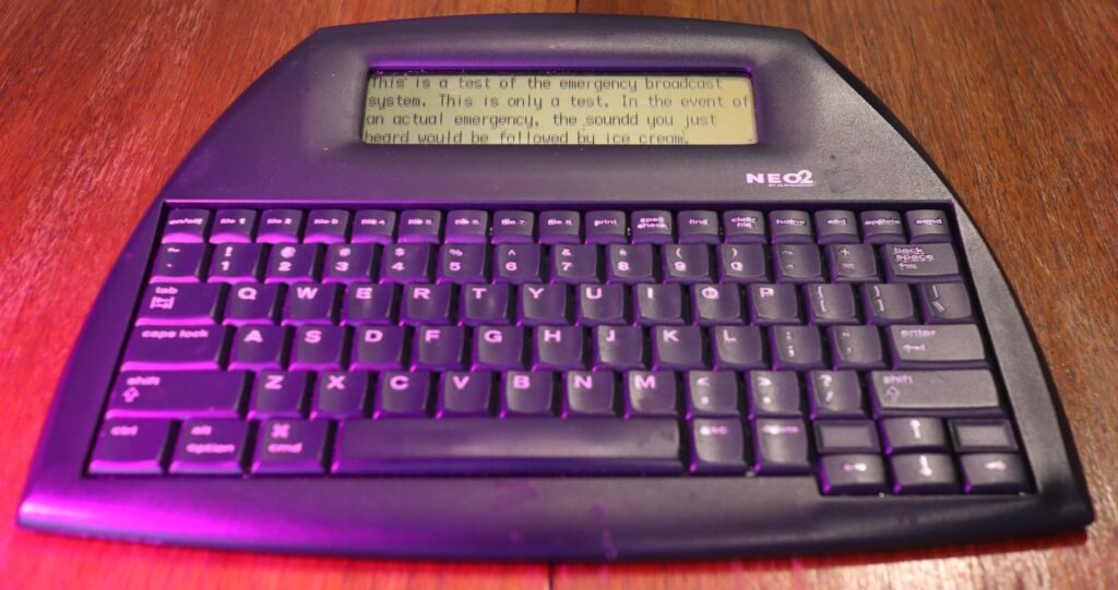 An AlphaSmart Neo2 device with a small screen at the top and a QWERTY keyboard below, seen here in a purple light.