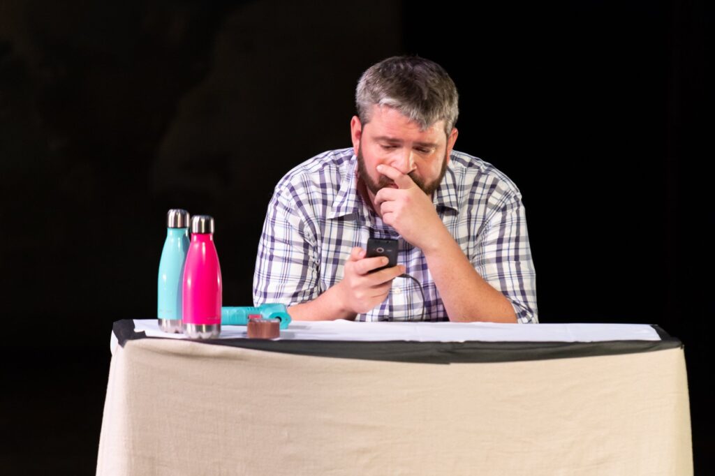 Actor in a plaid shirt at a table reading his phone, with colored water bottles at his side.