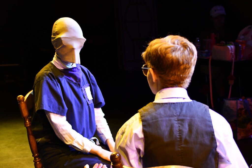 Two seated characters talking, The one facing the camera has his face covered with a piece of cloth that shows the shape of their glasses.