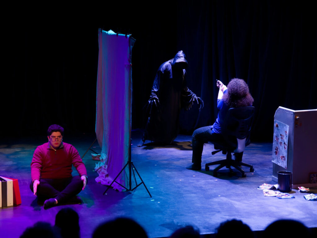 Photo of three seated characters on a stage, separated by a screen